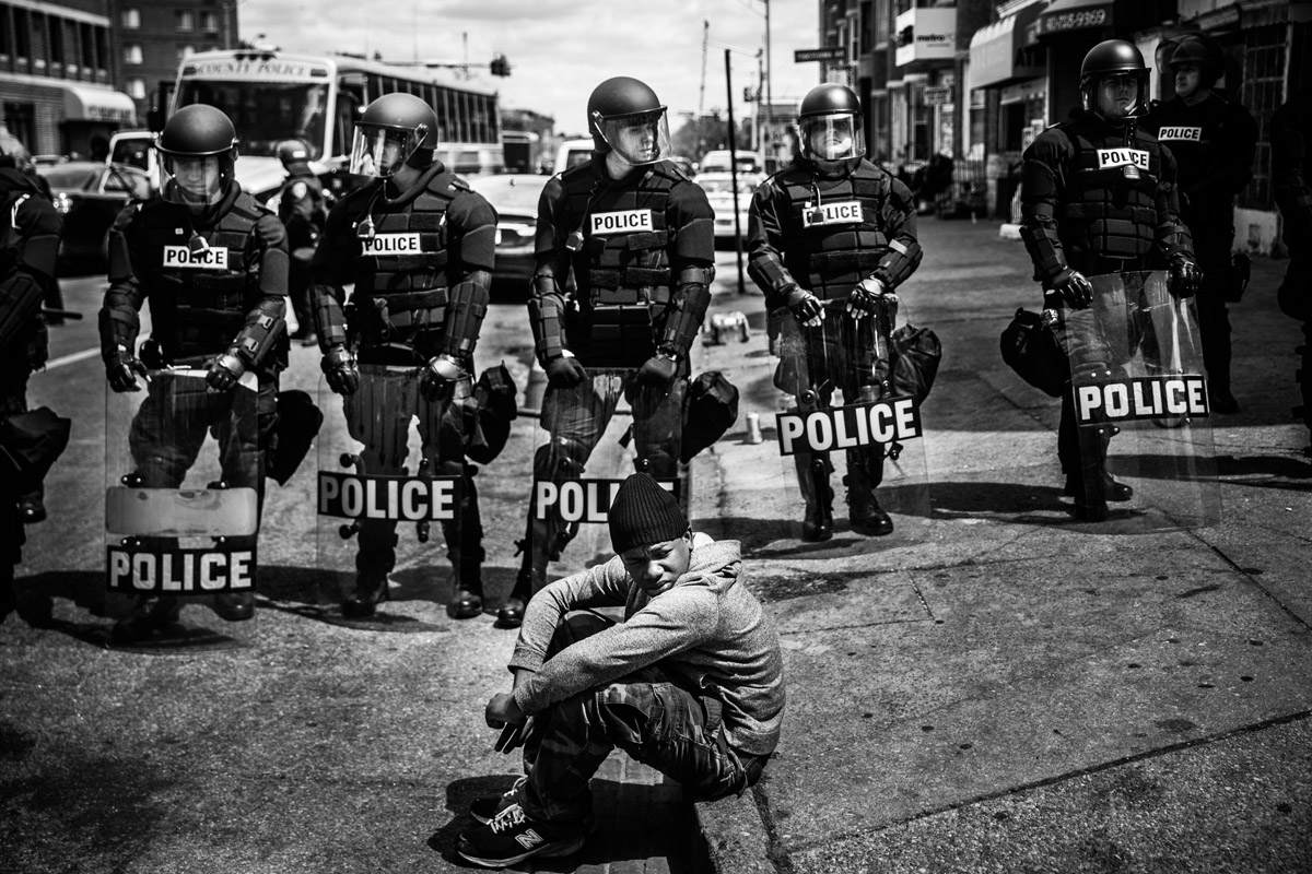 BALTIMORE, MD - APRIL 28: Daquan Green, age 17, sits on the curb while riot police stand guard near the CVS pharmacy that was set on fire yesterday during rioting after the funeral of Freddie Gray, on April 28, 2015 in Baltimore, Maryland.