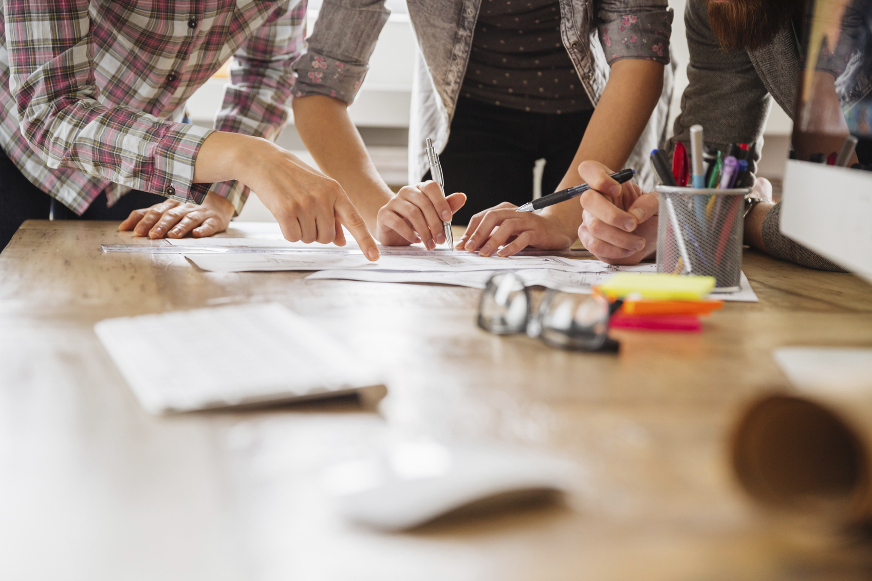 Picture of three pairs of arms and hands working at a desk. On the wooden table desk are also a coffee mug, glasses, post its and pens.