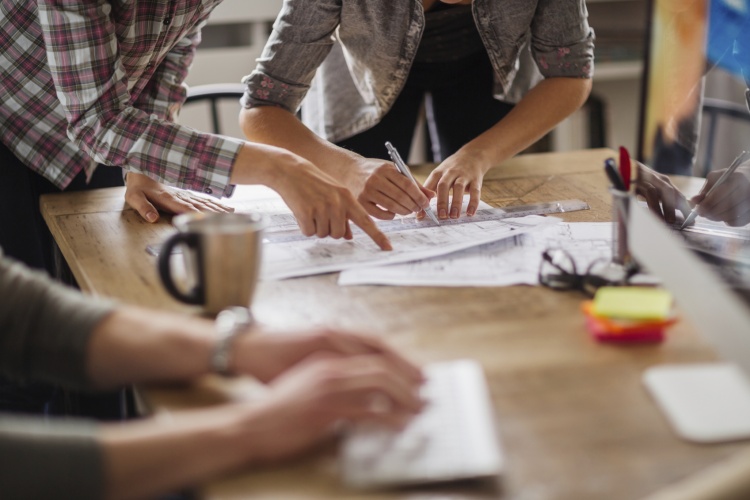 Picture of three pairs of arms and hands working at a desk. Two persons are working on a building design drawing on a paper and another one is typing on a keyboard in front of a computer screen. On the wooden table desk are also a coffee mug, glasses, post its and pens.