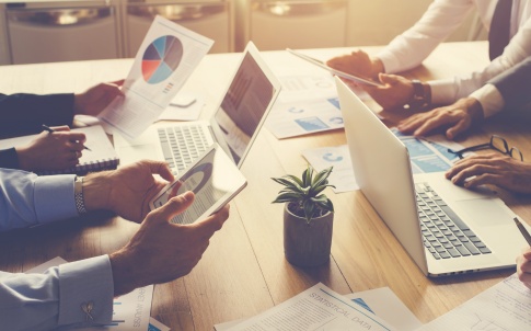 Group of people meeting with technology. They are around a conference table with digital tablets and laptops. There are marketing and strategy documents on the table