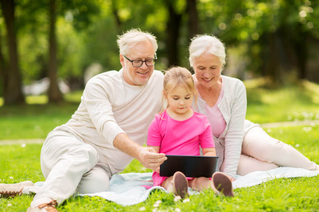 Frau und Mann sitzen mit Mädchen und Tablet-PC auf grüner Wiese.