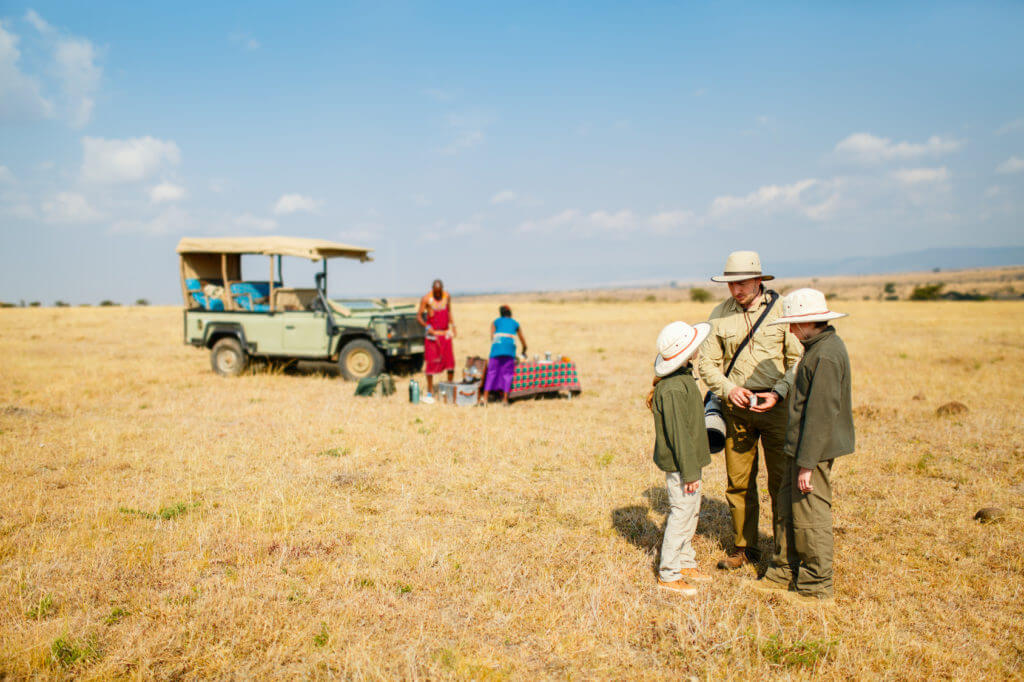 Touristen mit Safarihüten in der Savanne Afrikas, vor parkendem Fahrzeug.