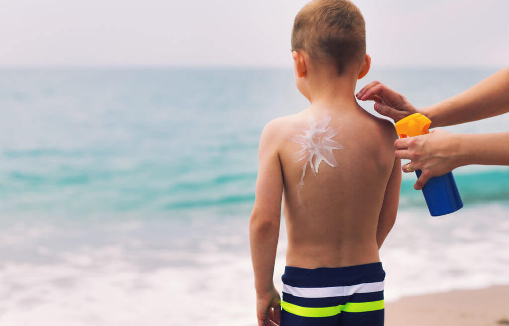 Frau cremt kleinen Jungen am Strand mit Sonnencreme ein.