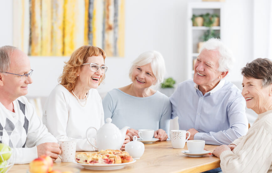 Senioren-Gruppe sitzt lächelnd bei Kaffee und Kuchen am Tisch.