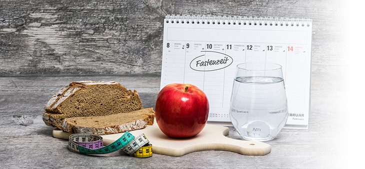 Brot, Apfel, Wasserglas und Maßband liegen vor einem Kalender, auf dem "Fastenzeit" steht.