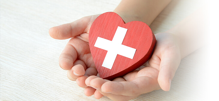 Hände halten rotes Herz aus Holz mit weißem Kreuz darauf.