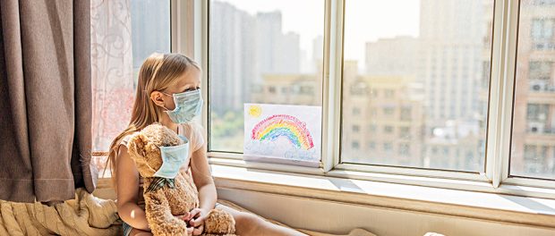 Kid with painted rainbow during Covid-19 quarantine at home. Caucasian Girl with teddy bear toy near window. Stay at home Social media campaign for coronavirus prevention, let's all be well