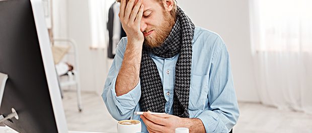 Sick bearded male in despair uses thermometer to measure temperature of body, sadly looks at it, suffers from high temperature, feels bad, surrounded by medicine and handkerchiefs at his workplace.