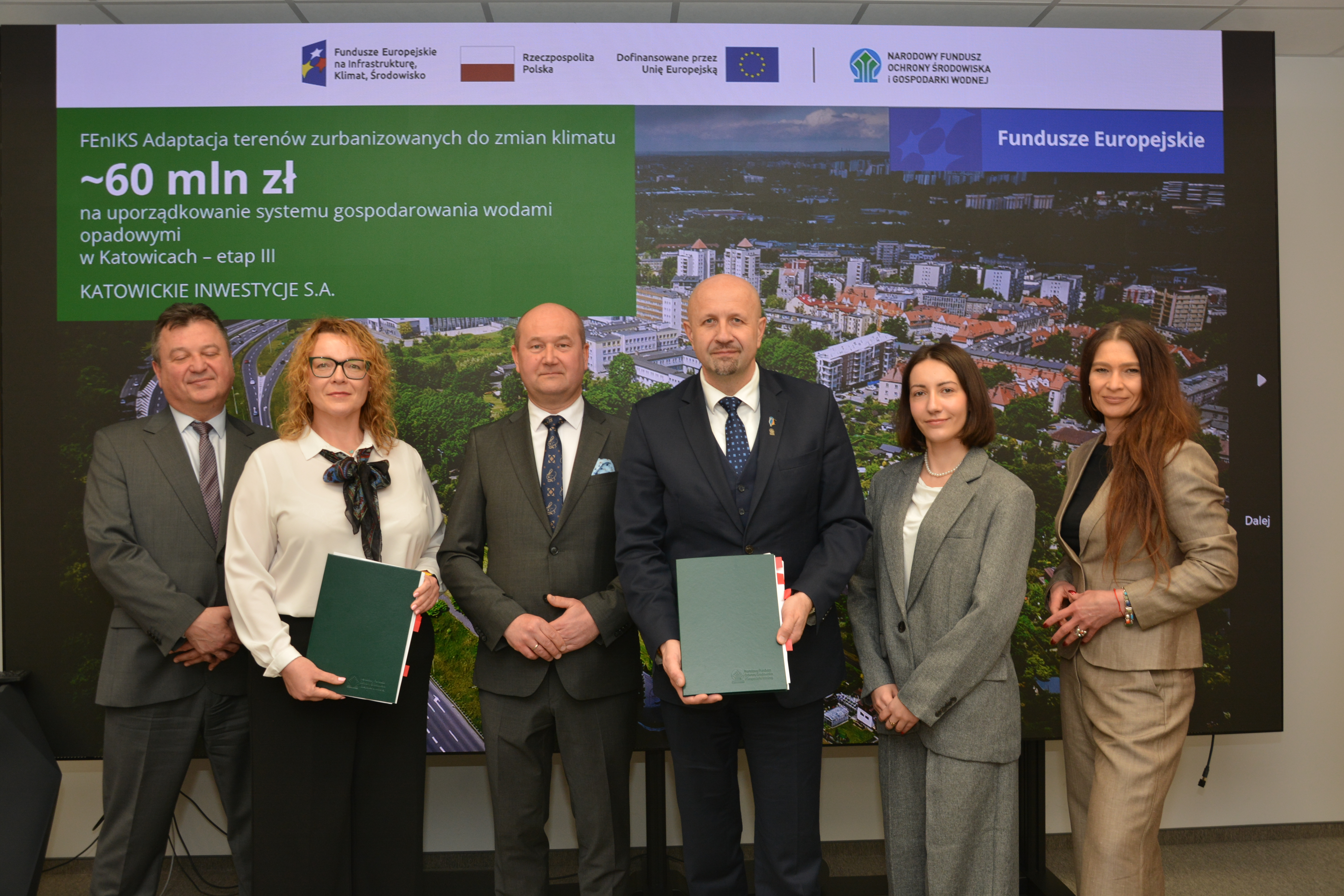 zdjęcie z podpisania umowy o dofinansowanie, obrazuje 6 osób stojących na tle planszy z widokiem miasta katowice i informacją o projekcie