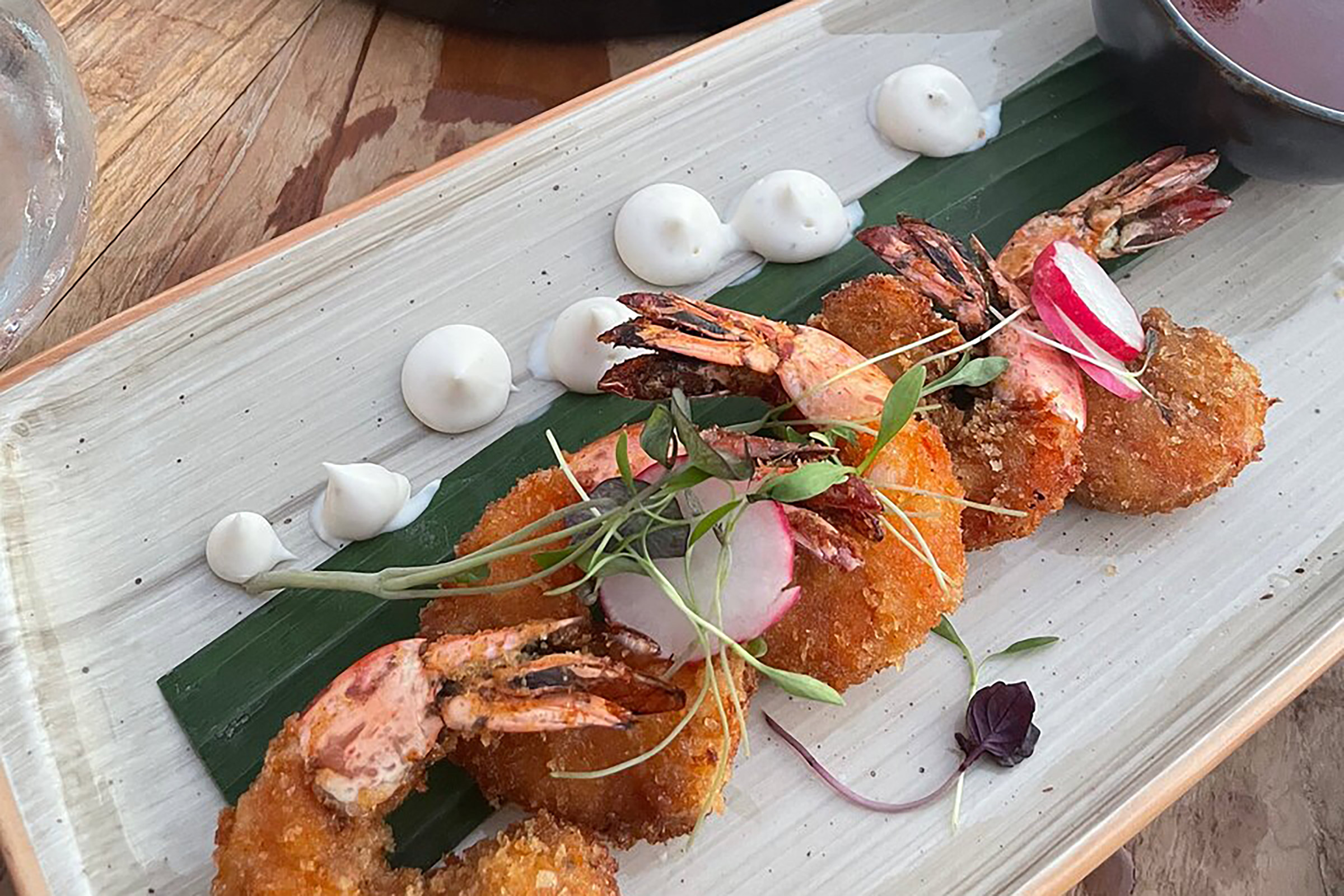 The extensive cocktail list boasts a wide collection of inspired signature recipes with wildflowers and fresh fruit lying in their core and adding the exotic touch anticipated in such a mesmerising setting, as well as the all-time classics expertly executed. The delicious dishes perfectly compliment the whole experience, as innovative tartars, crostinis, bao buns, burgers, premium meat cuts and many more delicacies give you something to nibble on as you enjoy the sun falling into the sea and the whitewashed houses of Mykonos Town painted in all shades of pink. - Secondary Image