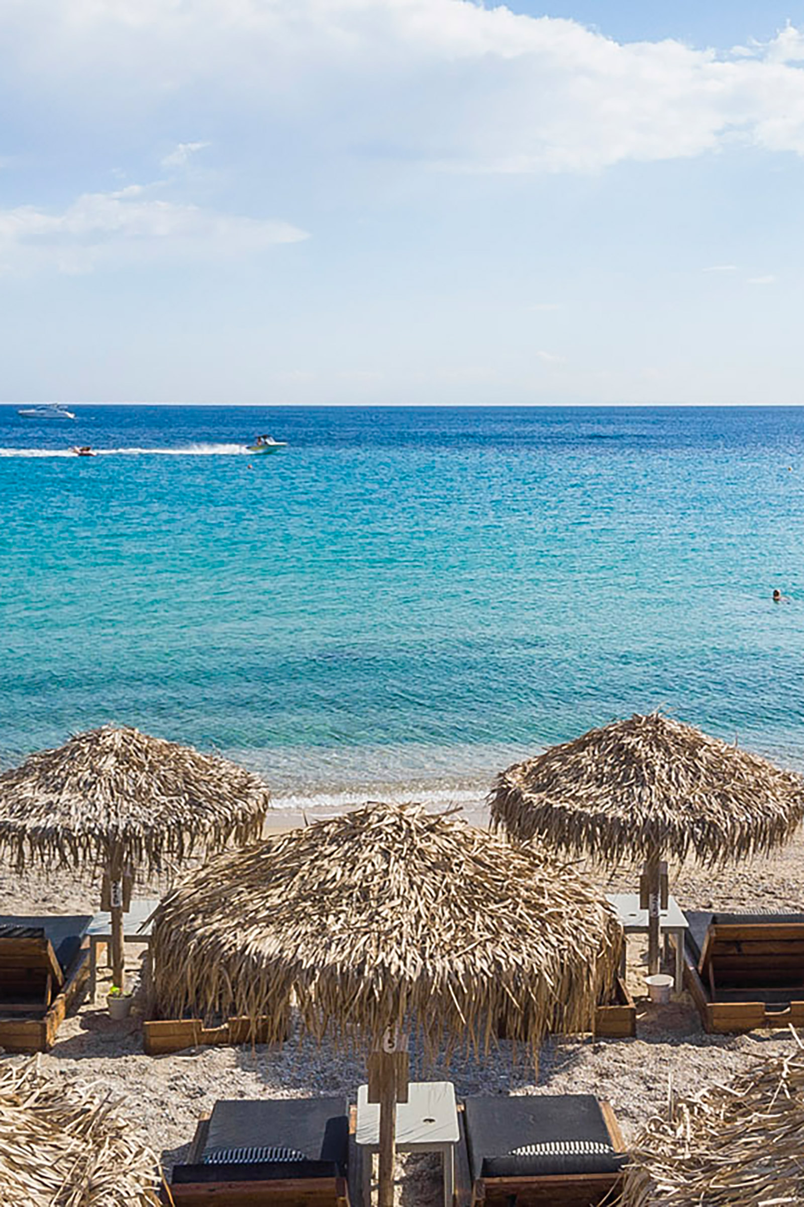 Relax all day on the golden sand of the pristine Elia beach, enjoy an innovative cocktail or a refreshing glass of wine, read your book, or just absorb the sun and the endless views, before heading for the robust wooden tables of the elegant restaurant awaiting to offer you a true feast for all the senses. - Image