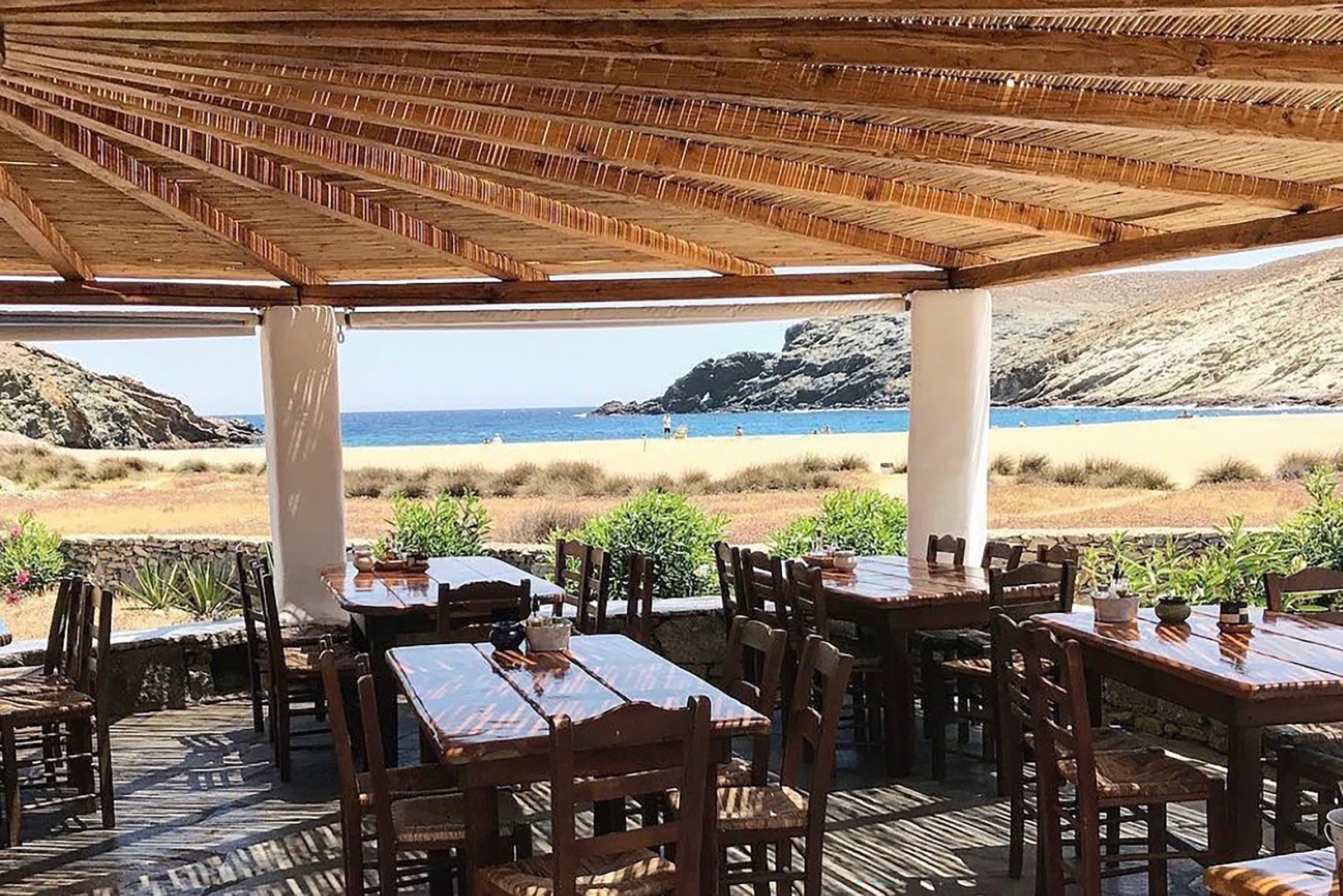 Far away from the loud and glamourous Mykonos party scene, here you will find yourself diving in and out the pristine waters all day long and then taking a seat at a perfect view table with the sand still between your toes and your hair still wet from the sea. - Secondary Image