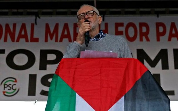 Jeremy Corbyn speaking at a lectern draped in a Palestinian flag.