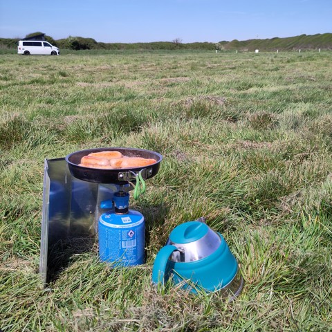 A large and still mostly empty camping field with a kettle and a camp stove frying This Isn't Pork Sausages in the foreground