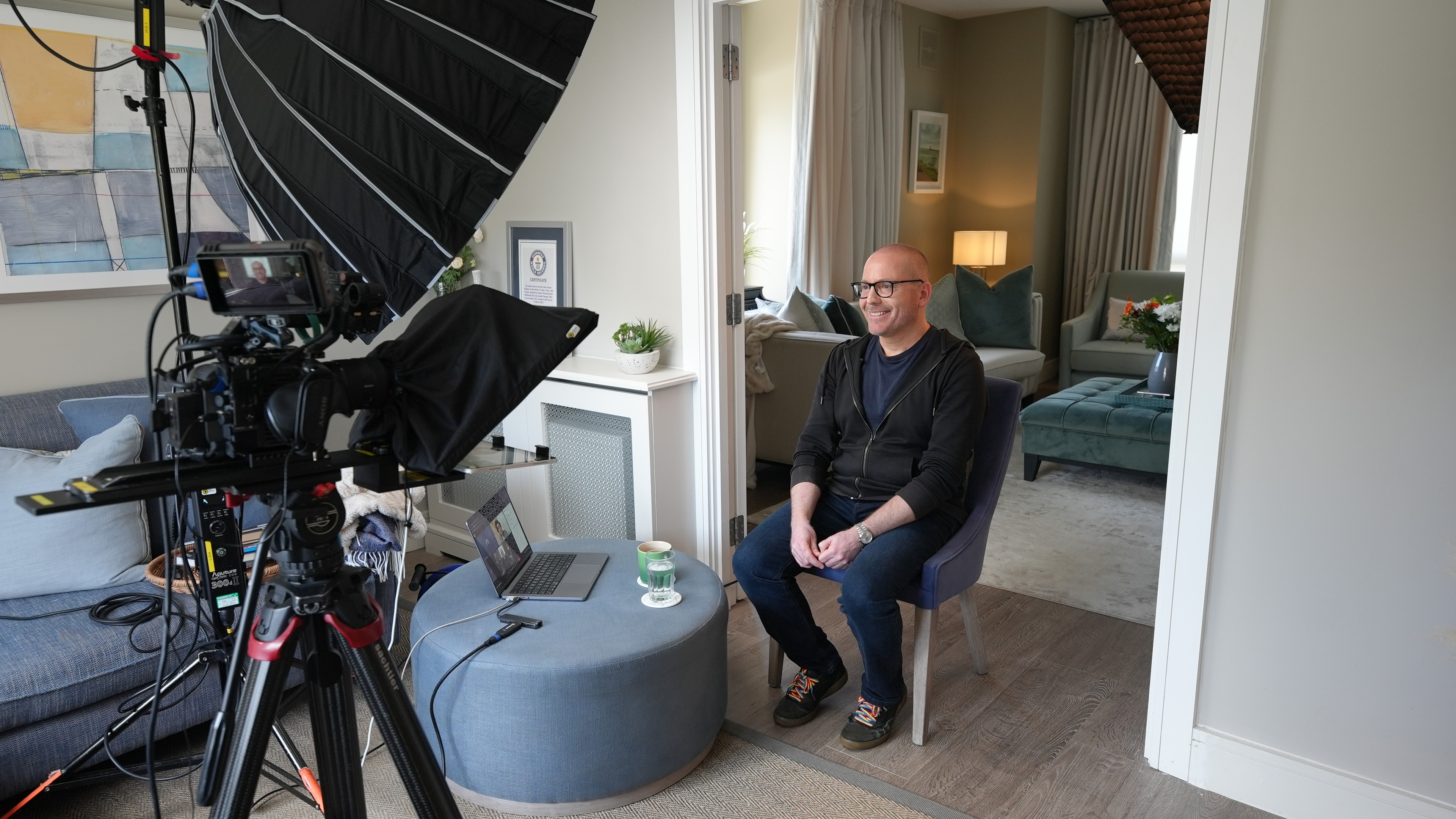 Shot of yours truly (white passing middle-aged guy with short hair and black glasses wearing a black hoodie, dark navy t-shirt, jeans, and shredded black skating sneakers with rainbow laces, sitting in front of a camera and light by a doorway.