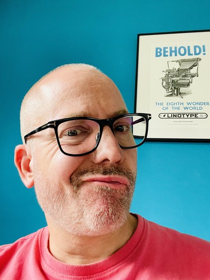 A photo of yours truly wearing a pink t-shirt and pulling a face with a poster behind him with an etching of a Linotype machine and LINOTYPE written in blank ink and “Behold! The eight wonder of the word” in blue.