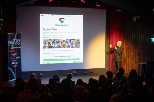 Me (white passing guy with short hair wearing a brown tweed jacket, black t-shirt, and jeans), presenting to a small audience in front of a screen that has the Gaza Verified web site on it.