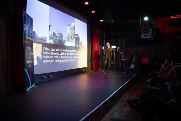 Me (white passing guy with short hair wearing a brown tweed jacket, black t-shirt, and jeans), presenting to a small audience in front of a screen that has Baraa’s photo and a quote by her saying “You can see us loading our belongings on to a truck, leaving behind the home I grew up in. With the help of @aral and Joy, my family … managed to evacuate to the South” (bits of the text are blown out and illegible due to glare from a spotlight.