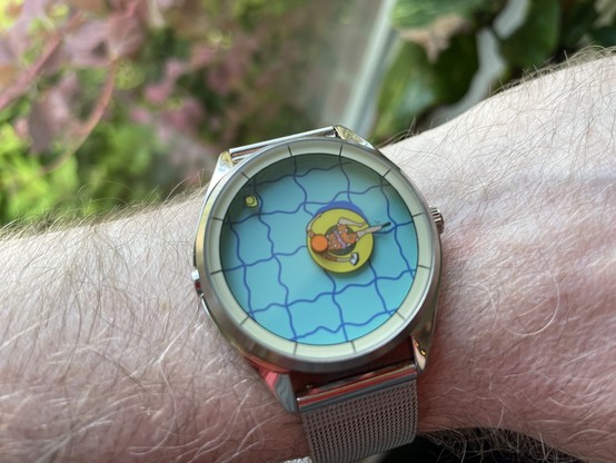 A simple metallic quartz watch with a hand-painted illustration of a person floating on a yellow rubber ring in light blue pool with the pool lines distorted by the water and yet lining up with the tiles at the side that make up the hour markers. One of their legs is sticking out, pointing out the hour and a little rubber ducky that circles the circumference is the minute hand. The illustration is by Kristof Devos.
