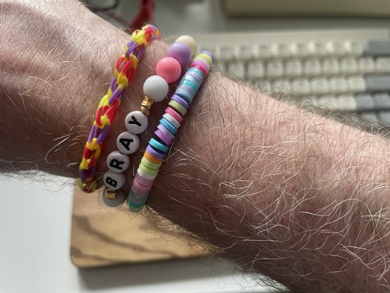 A rather thin, pale, and hairy arm with three hand-made multicolour plastic bracelets.
One has little chains, the other beads and the letters B R A Y and the last litte rings.