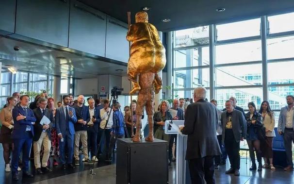 The Orange Plague: view from back of statue on display at EU Parliament of gold-plated naked Trump holding golf club on the shoulders of a thin man. People stand around it as someone addresses them from behind a podium.
