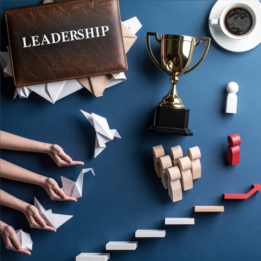 Es werden Gegenstände dargestellt die mit Leadership in Verbindung gebracht werden. Ein Pokal, eine Kaffeetasse, Bausteine und Holzfiguren wovon eine rot ist und die Führungsrolle symbolisiert