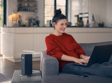 Frau sitzt auf Couch mit Laptop und neben ihr steht ein Router