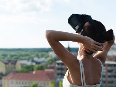 Eine Frau mit VR-Headset steht auf dem Dach und legt die Arme in den Kopf, im Hintergrund sieht man Häuser und Dächer einer Stadt.