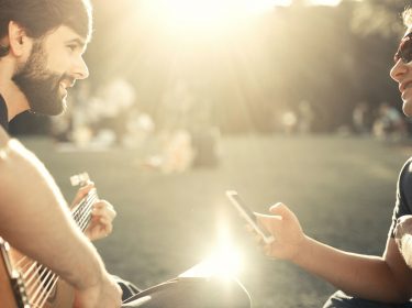 Männer sitzen im Park mit Gitarre und Smartphone.