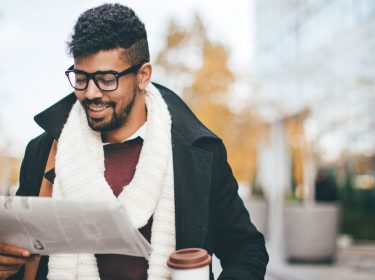 Junger Mann liest Zeitung und hält Kaffeebecher in der Hand.