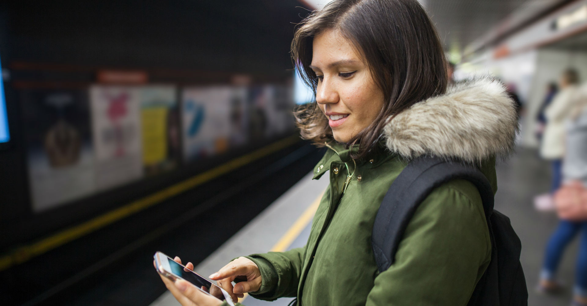 Lächelnde Frau am Bahnsteig schaut auf ihr Smartphone.