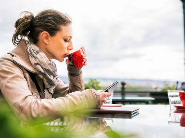 Frau trinkt Kaffee aus einer roten Tasse und schaut auf ihr Smartphone.