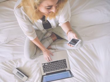 Lachende Frau auf Bett mit iPhone und MacBook.