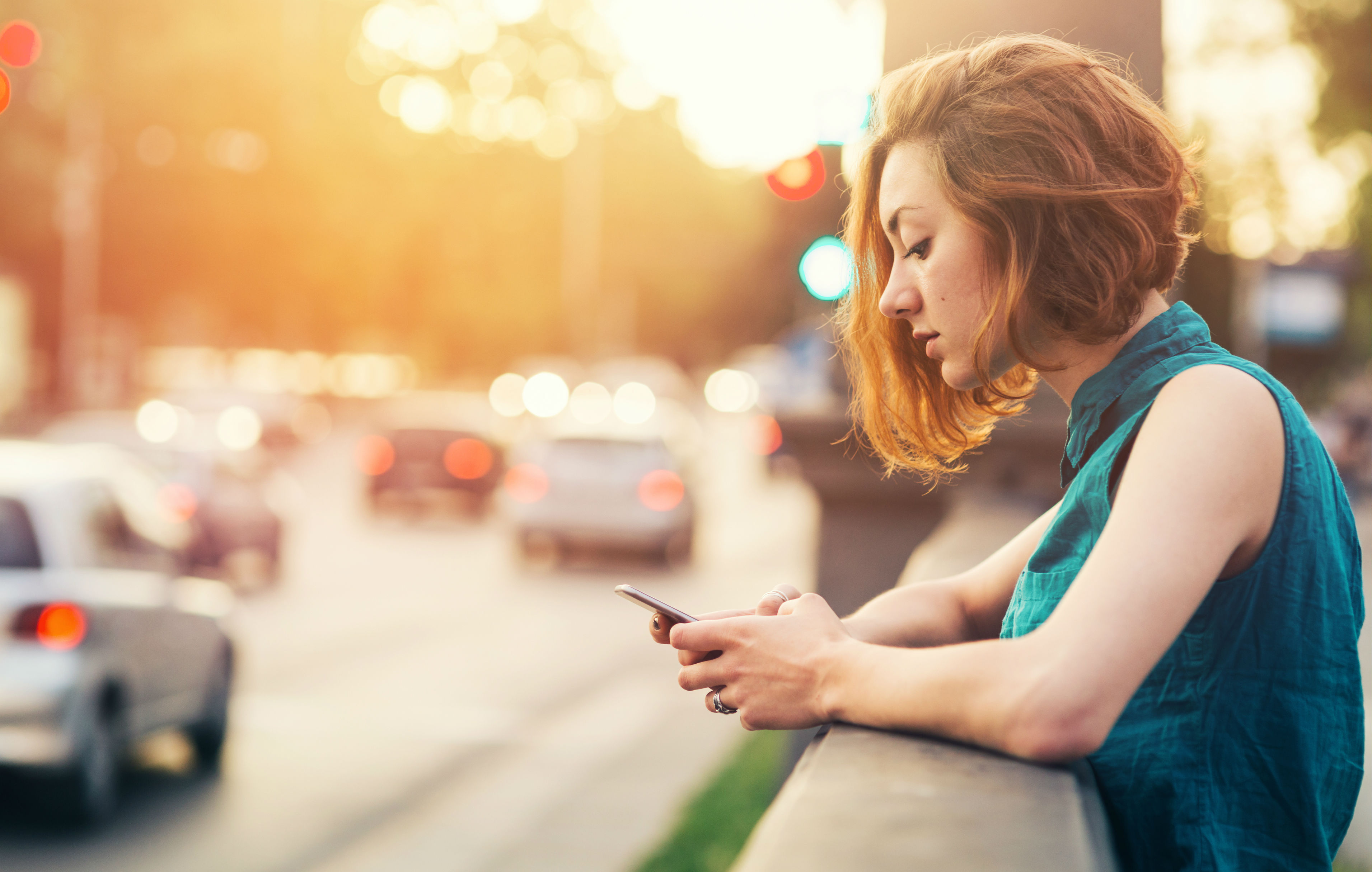 Junge Frau mit roten Haaren lehnt sich an eine Mauer und schaut auf ihr Smartphone.