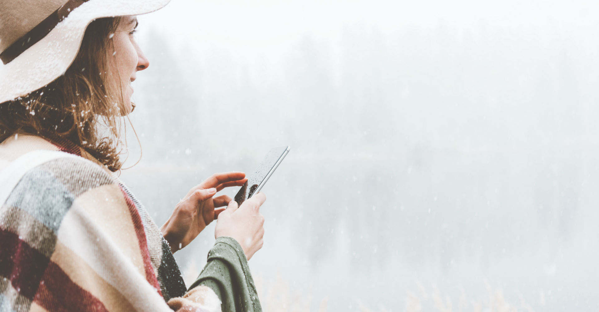 Junge Frau steht lächelnd im Schnee und hält dabei ein Smartphone in der Hand.