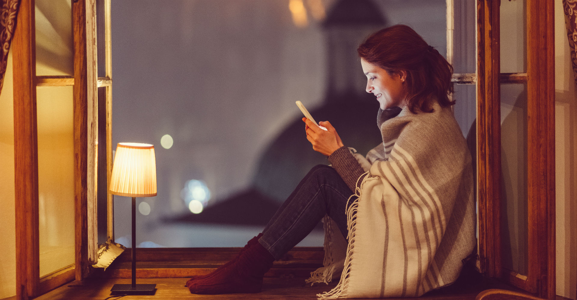 Junge Frau sitzt am Fenster mit ihrem Smartphone und schreibt eine Nachricht.