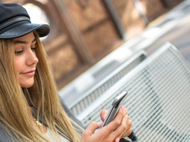 Junge Frau sucht an einer Bushaltestelle nach Fernbus-Reisen via Smartphone