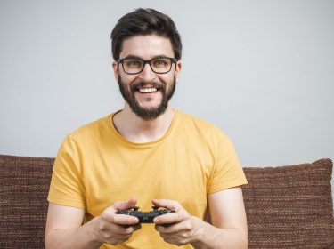 Junger Mann sitzt auf dem Sofa und zockt via Controller Assassin's Creed Origins.