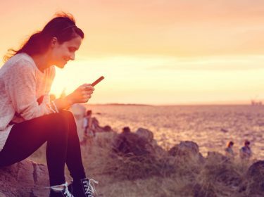 Junge Frau sitzt mit ihrem Smartphone am Strand.