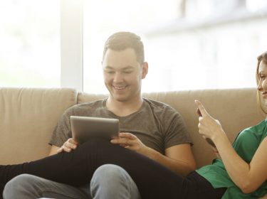 Junges Paar entspannt mit Smartphone und Tablet auf der Couch.