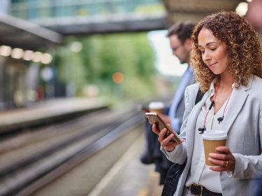 Eine junge Frau steht am Bahngleis und schaut auf ihr Smartphone.