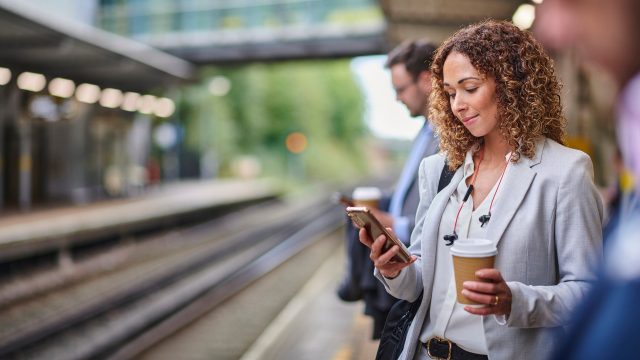 Eine junge Frau steht am Bahngleis und schaut auf ihr Smartphone.
