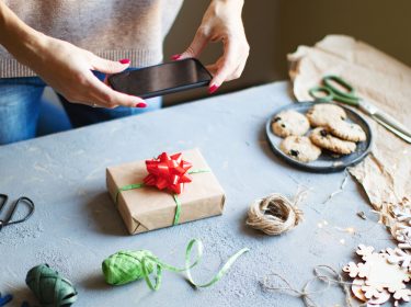 Eine Frau fotografiert ein von ihr verpacktes Geschenk mit dem Smartphone auf einem Basteltisch.