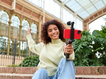 Eine junge Frau sitzt auf Steinstufen und hält einen Selfiestick mit Smartphone in der Hand.