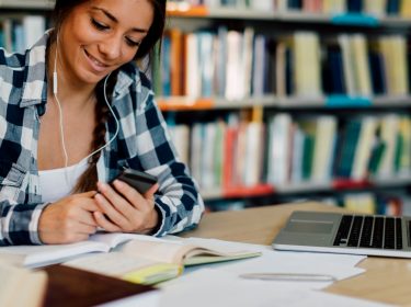 Junge Frau in Bibliothek lernt mit Laptop und am Smartphone