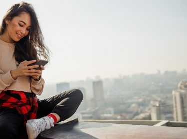 Frau sitzt auf einer Mauer und schaut auf ihr Handy.