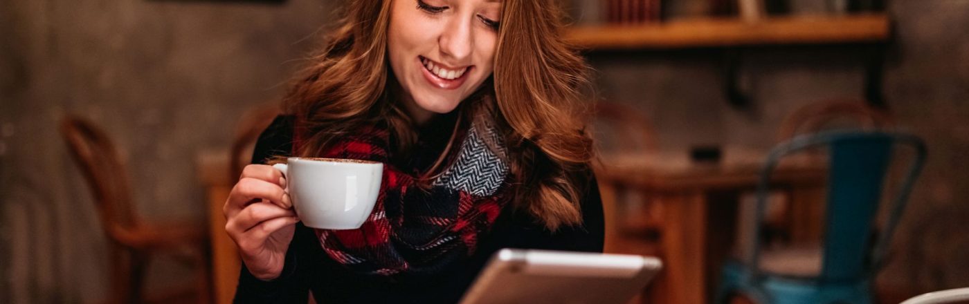 Junge Frau sitzt mit einem Tablet und einer Tasse Kaffee in der Hand auf einem Stuhl