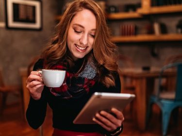 Junge Frau sitzt mit einem Tablet und einer Tasse Kaffee in der Hand auf einem Stuhl