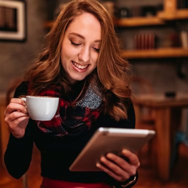 Junge Frau sitzt mit einem Tablet und einer Tasse Kaffee in der Hand auf einem Stuhl