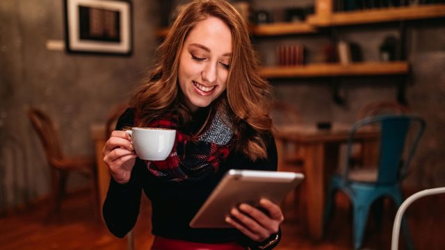 Junge Frau sitzt mit einem Tablet und einer Tasse Kaffee in der Hand auf einem Stuhl